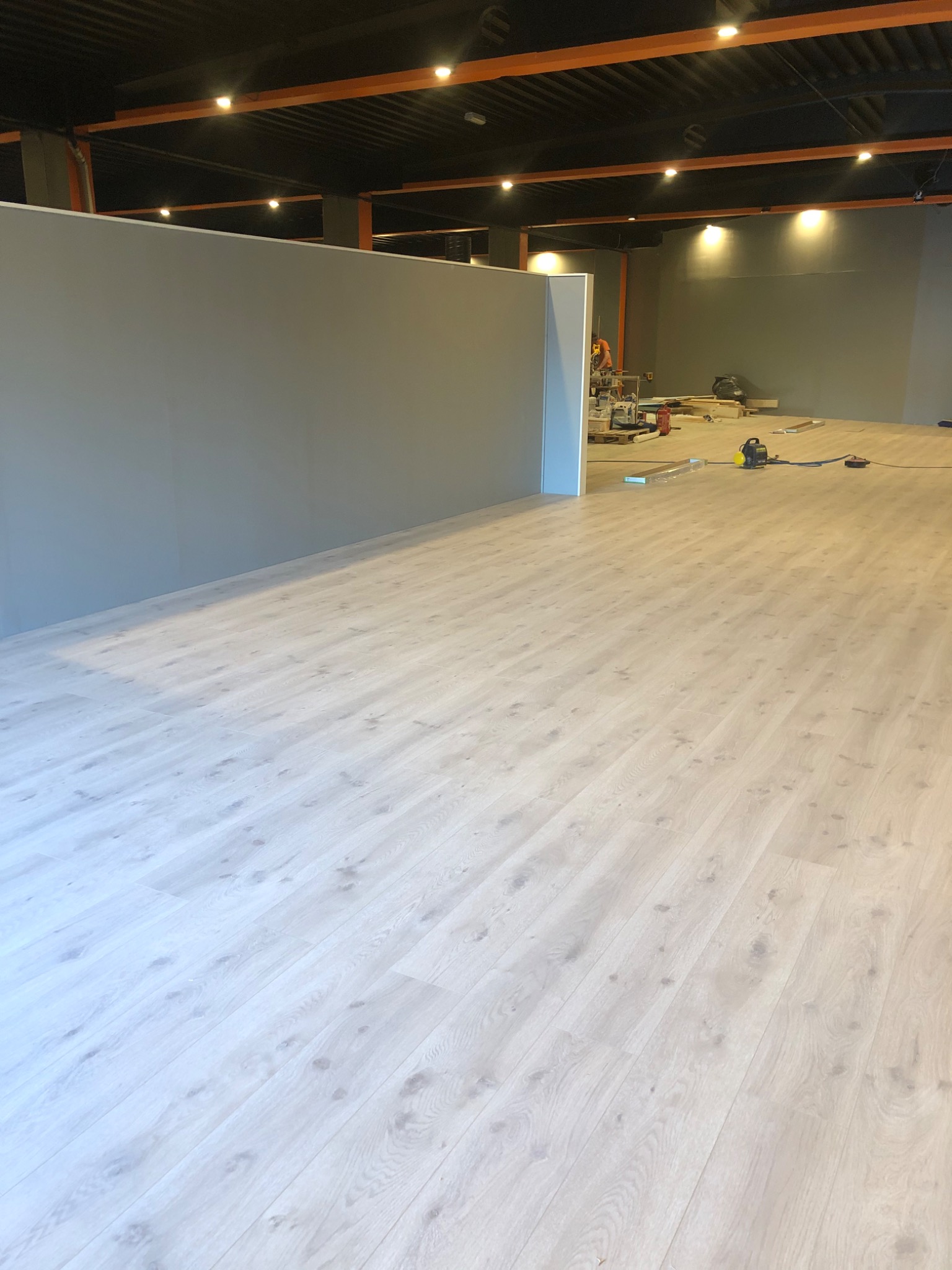 Jasne panele podłogowe ułożone w nowym pomieszczeniu, widoczne narzędzia i materiały budowlane w tle, ściany w odcieniach szarości, czarne oświetlenie sufitowe.