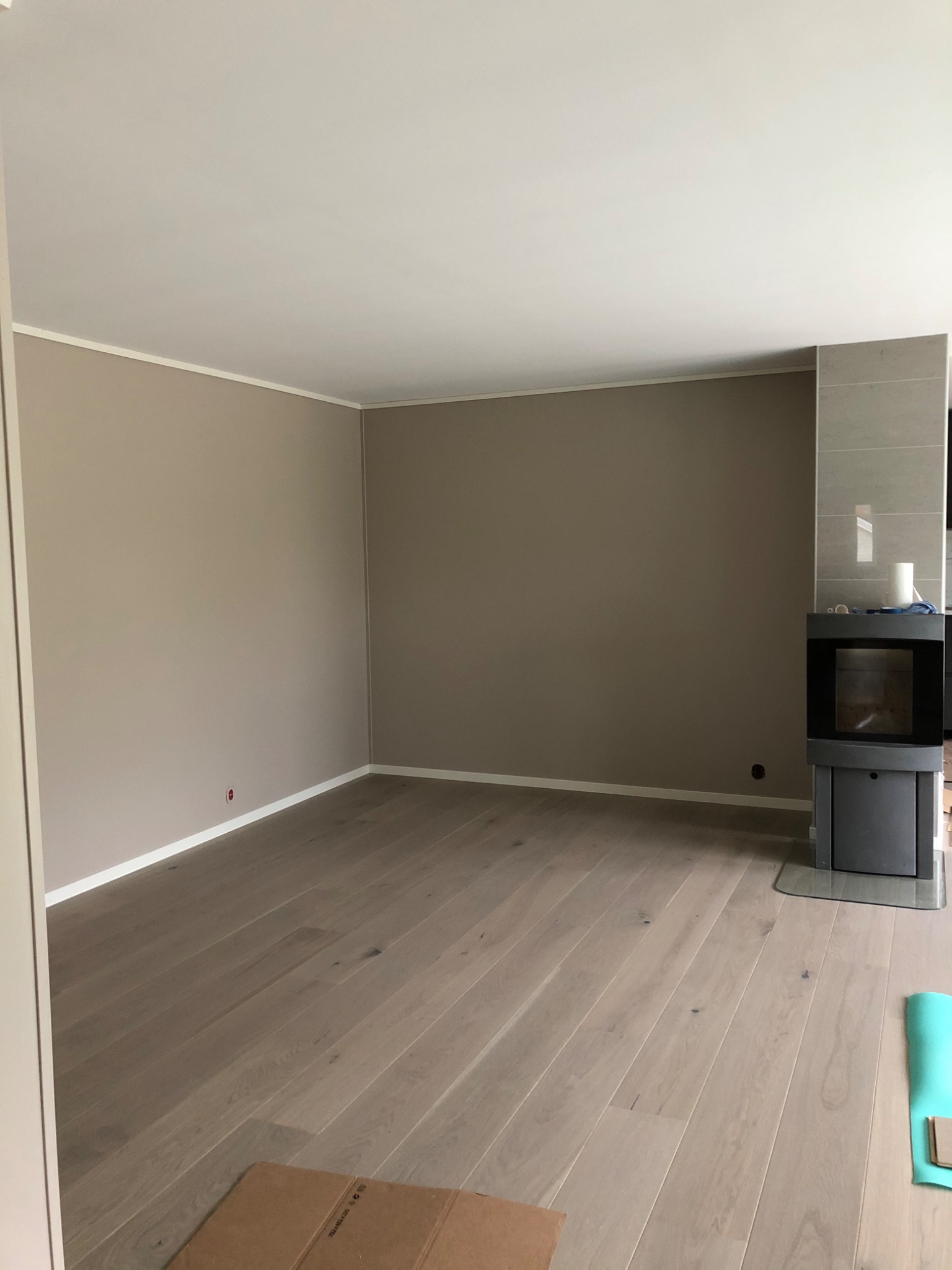 Pomalowane na beżowo wnętrze pokoju z jasną podłogą z paneli i nowoczesnym, szarym kominkiem narożnym. Widoczny fragment kartonu na podłodze i mata do ćwiczeń. Świeżo pomalowane ściany z białymi...