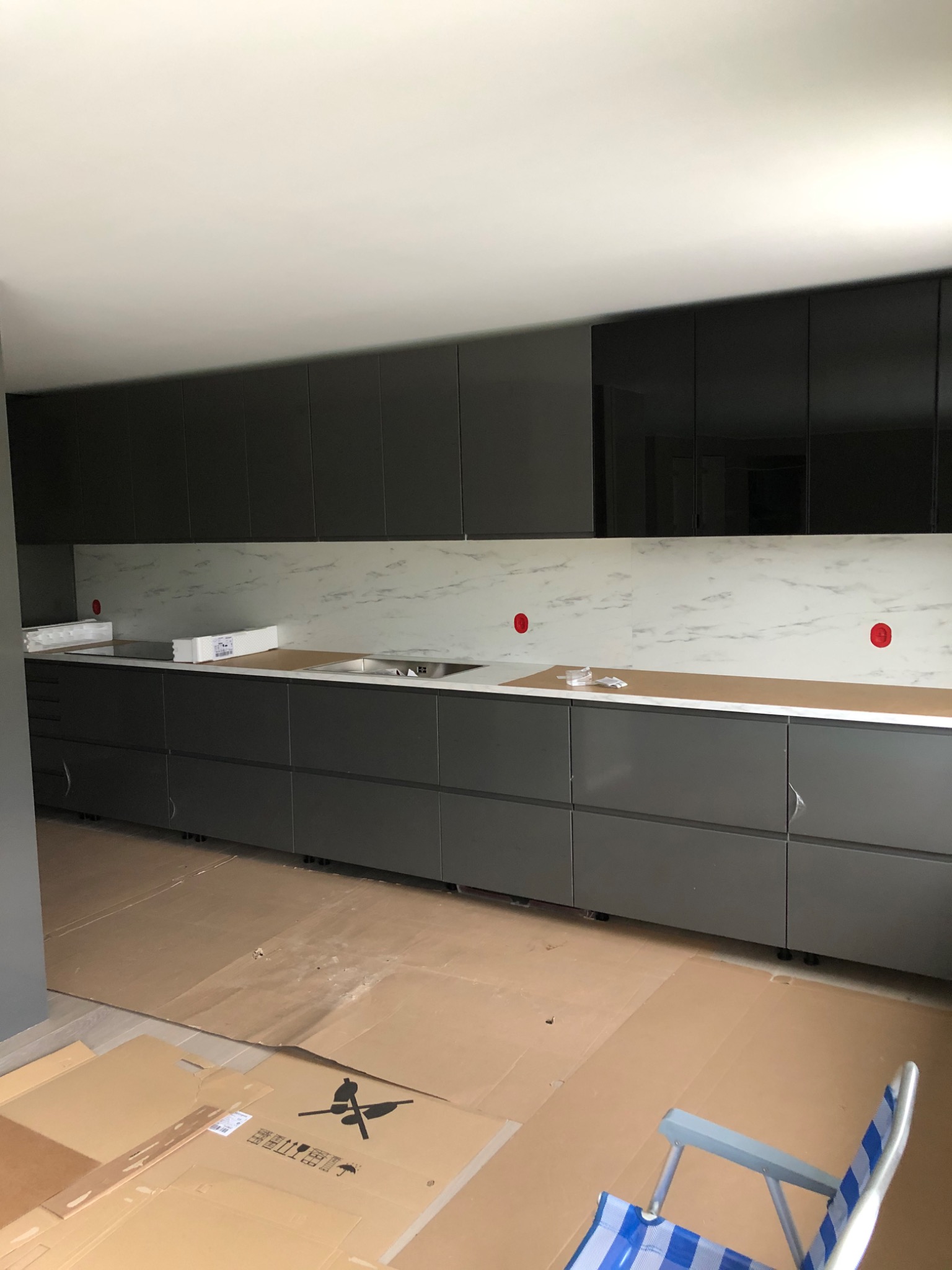Nowoczesna kuchnia w trakcie montażu: szare szafki z połyskiem, biały blat z marmurowym wzorem, podłoga zabezpieczona kartonem, krzesło plażowe w rogu.