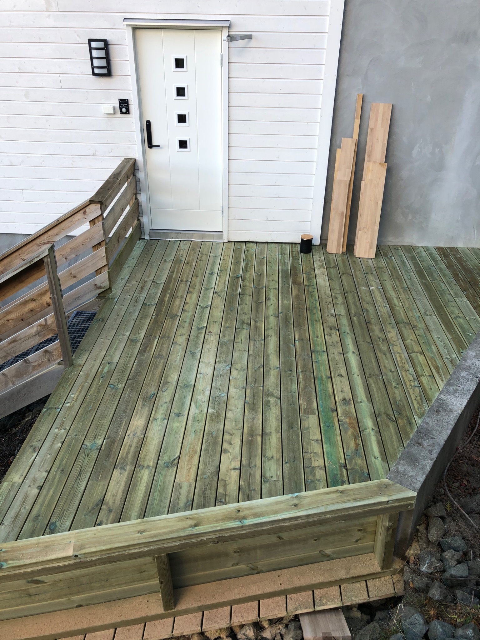 Drewniany taras przed wejściem do domu, z widocznymi deskami podłogowymi w odcieniach zieleni i brązu, oraz fragmentem schodów z drewnianą barierką po lewej stronie i przygotowanymi deskami obok...