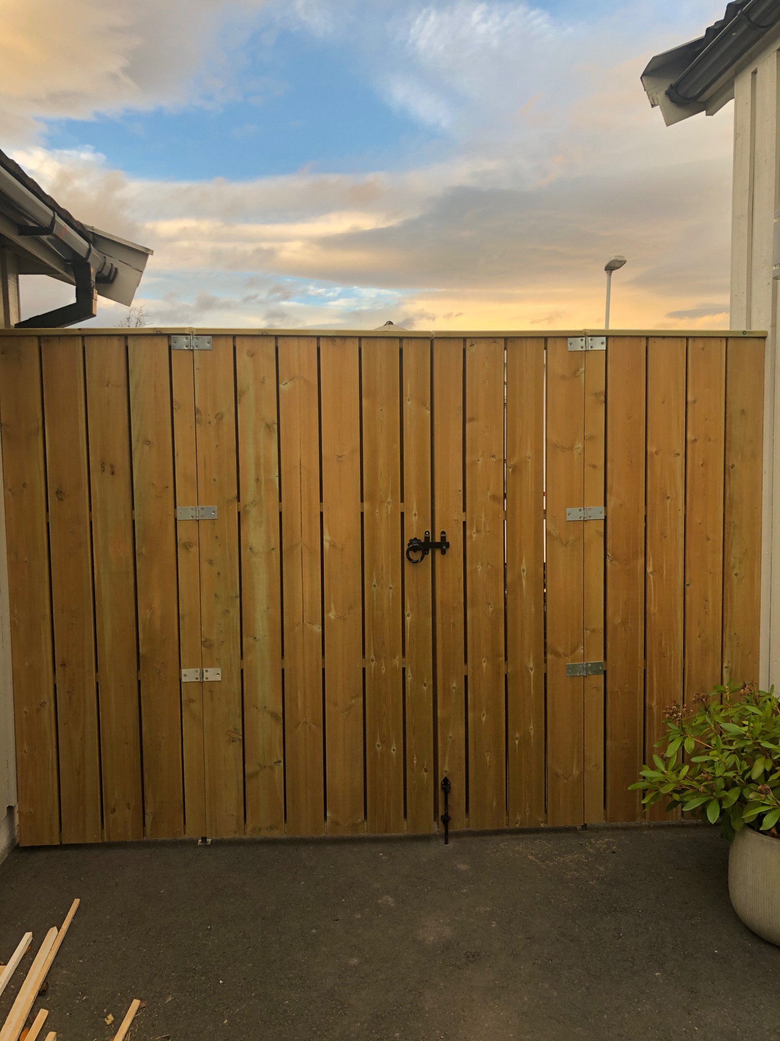 Nowa drewniana brama wejściowa z metalowymi zawiasami i czarnym zamkiem, widok z zewnątrz na tle błękitnego nieba z chmurami o zachodzie słońca.