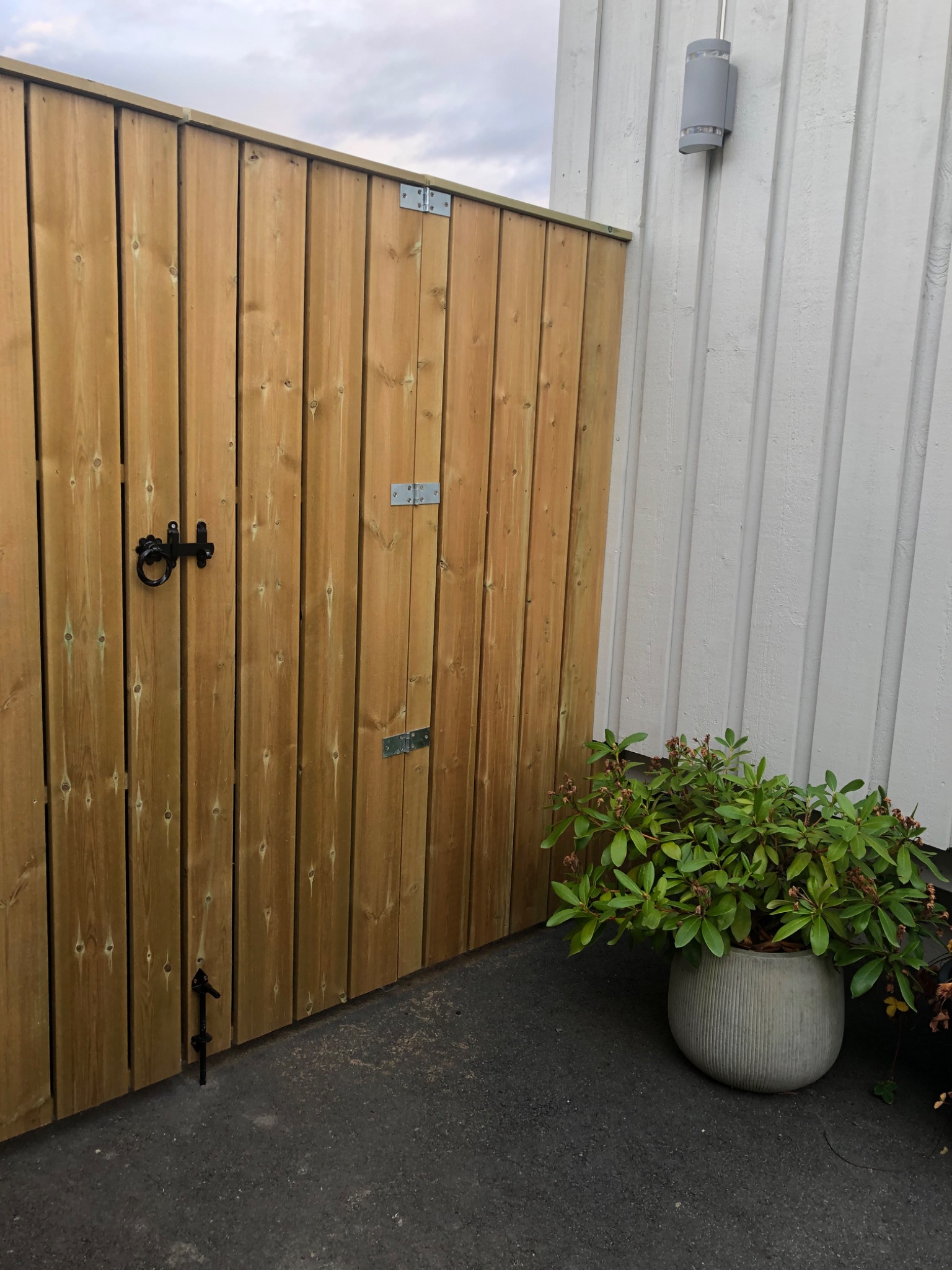 Drewniana furtka z metalowymi zawiasami i ozdobnym zamkiem, przylegająca do ściany budynku z lampą zewnętrzną, obok donica z zieloną rośliną.