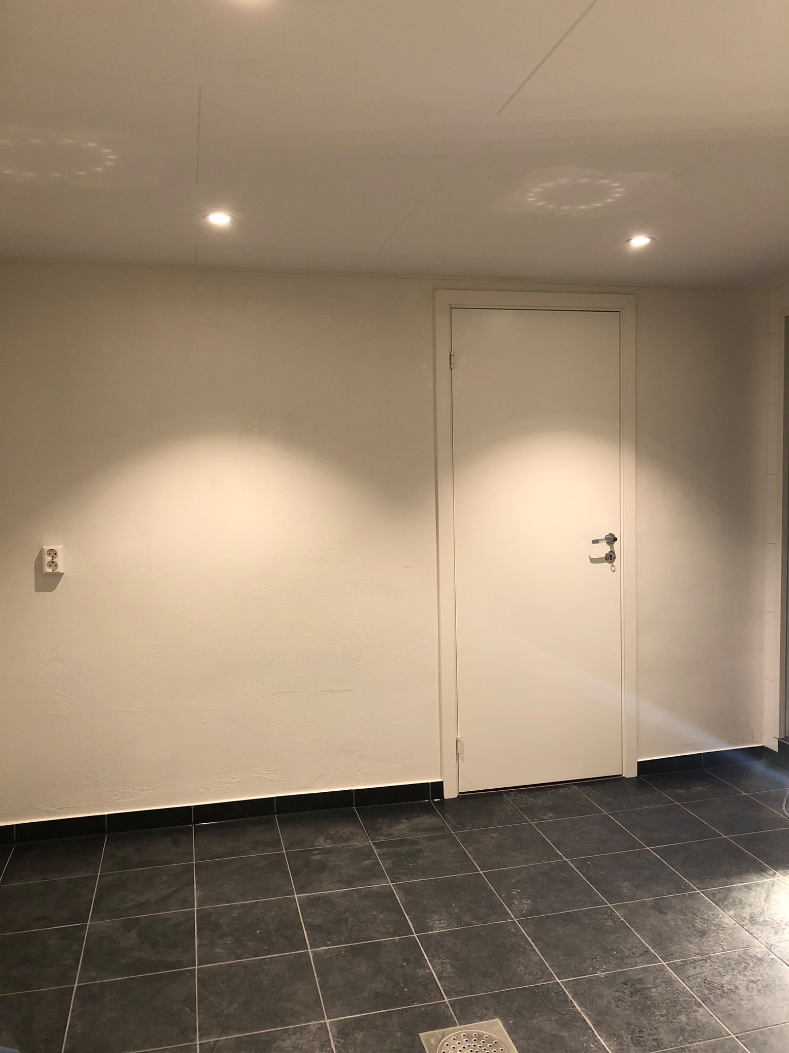Wnętrze po remoncie: Ściana w odcieniach bieli z widocznym gniazdkiem elektrycznym, białe drzwi z metalową klamką, czarna podłoga wyłożona kwadratowymi płytkami, oświetlenie sufitowe punktowe.