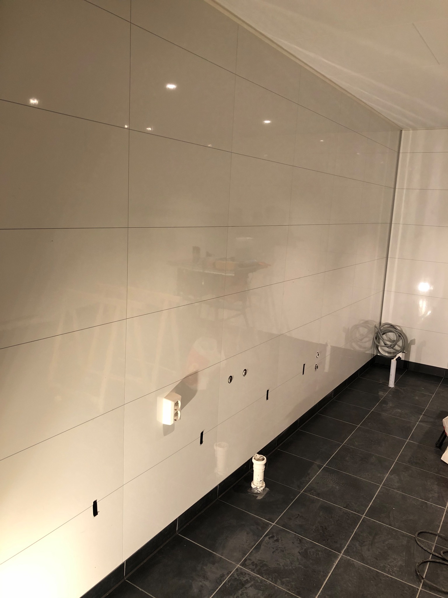 Błyszczące, kremowe płytki ceramiczne ułożone na ścianie łazienki, z widocznymi otworami na instalacje i gniazdko elektryczne; ciemna, kwadratowa podłoga z płytek i zwinięty kabel na podłodze.