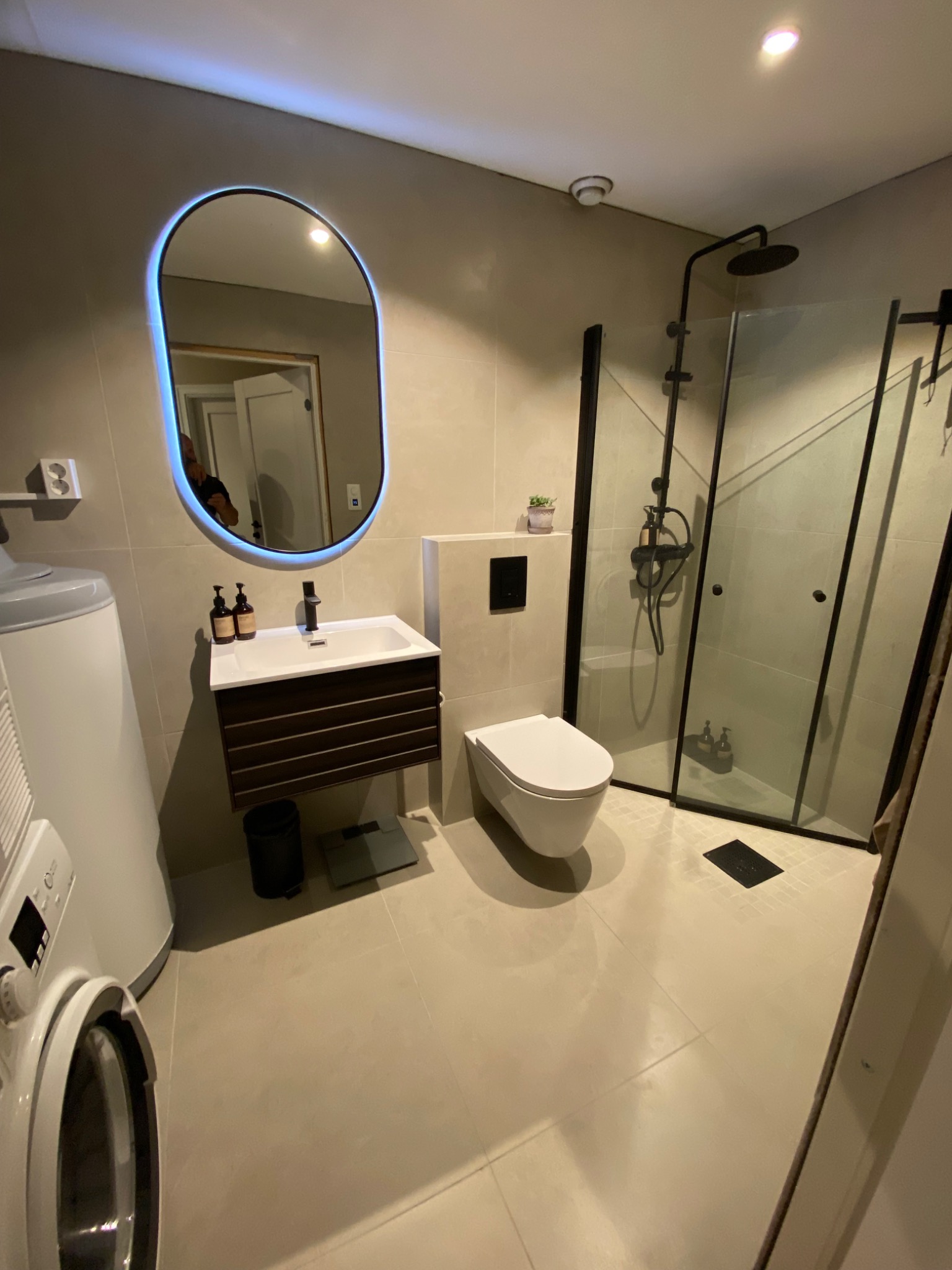 Nowoczesna łazienka z podświetlanym lustrem owalnym, kabiną prysznicową typu walk-in z czarnymi profilami, umywalką z szafką w kolorze ciemnego drewna i pralką na pierwszym planie.