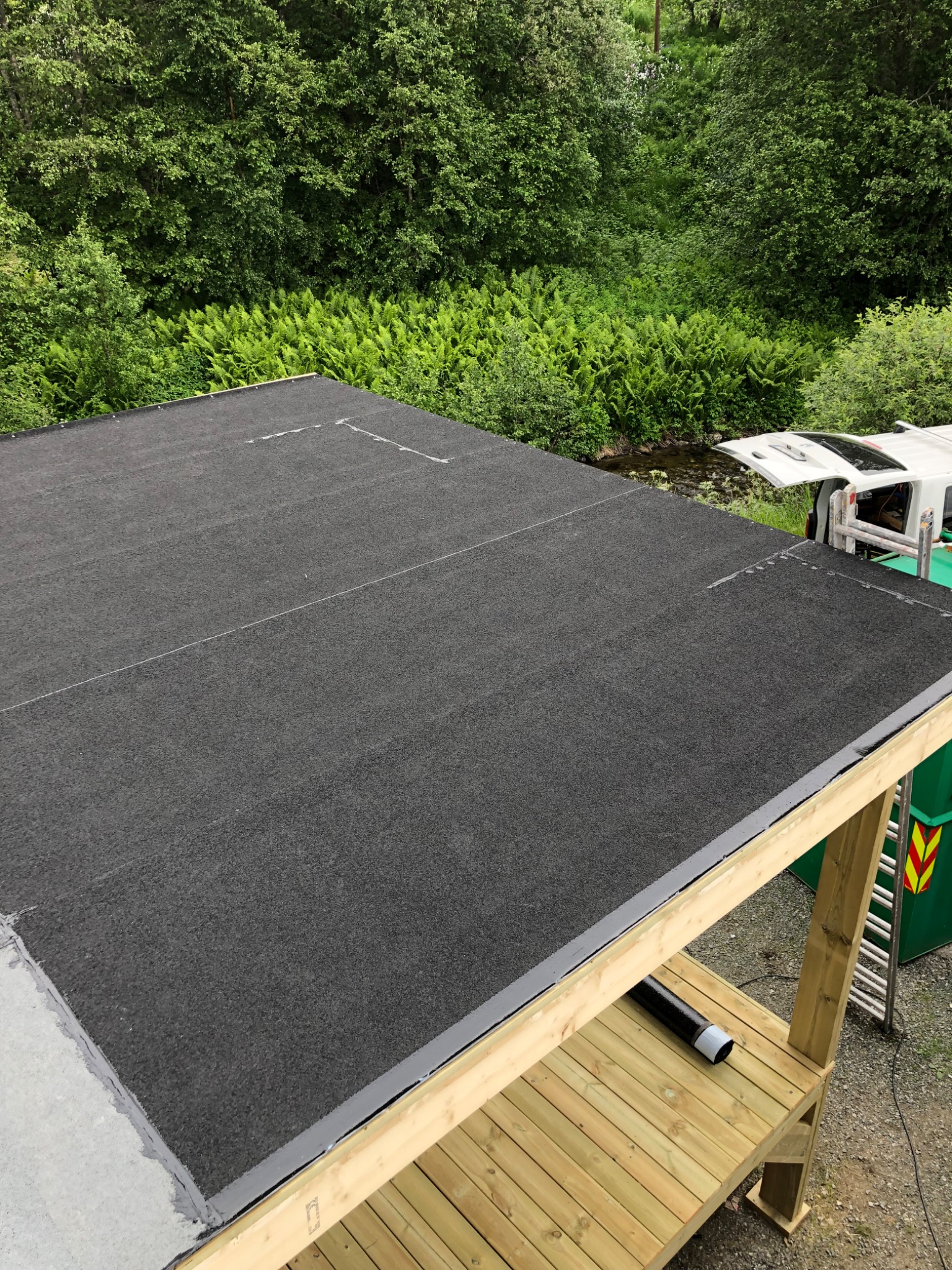 Czarna papa na dachu z widocznymi liniami łączeń, Koszalin, w tle bujna roślinność i częściowo widoczny samochód dostawczy.