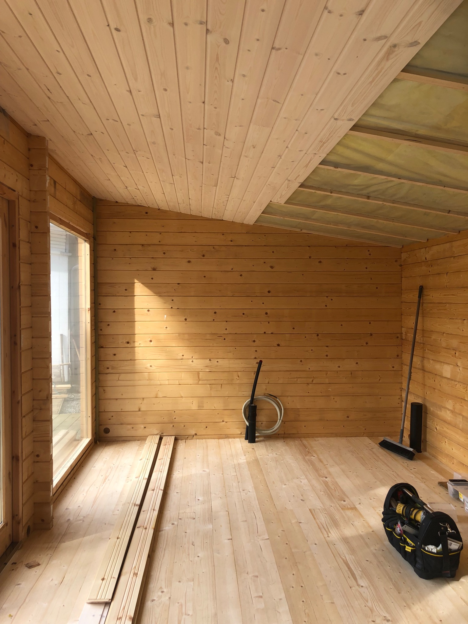 Wnętrze drewnianego domku z boazerią na ścianach i suficie, częściowo ocieplonym, z oknem i narzędziami w torbie na podłodze.