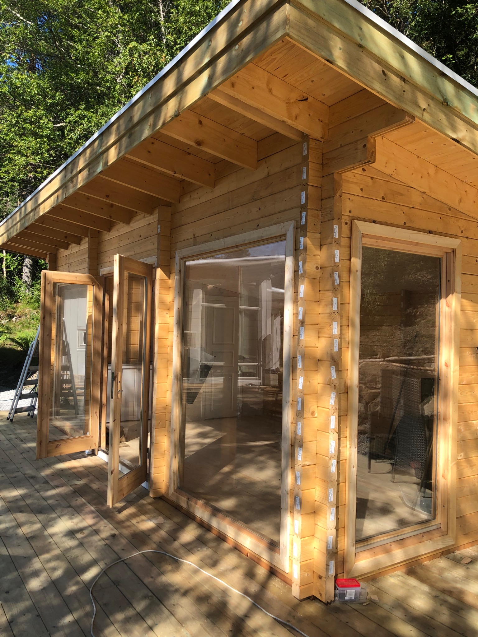 Drewniany domek letniskowy w trakcie montażu, widoczne otwarte przeszklone drzwi, duże okna i drabina oparta o ścianę, elementy konstrukcyjne z numerowanymi etykietami.