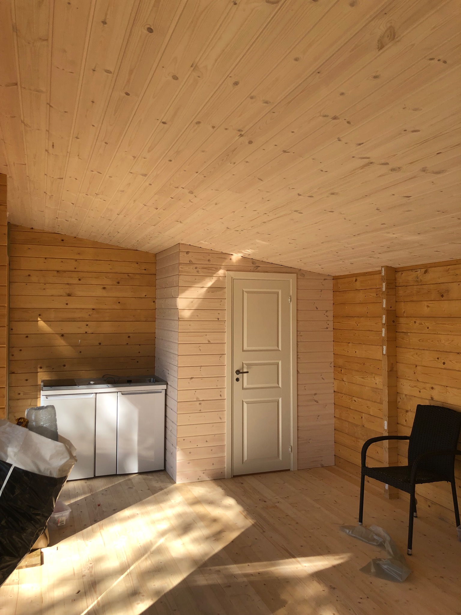 Wnętrze pomieszczenia wykończonego boazerią w jasnym odcieniu drewna, z białą zabudową kuchenną i czarnym krzesłem, wpadające promienie słoneczne tworzą długie cienie na podłodze.