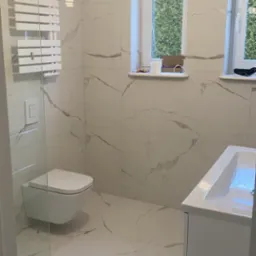 Nowoczesna łazienka z białymi płytkami imitującymi marmur, podwieszana toaleta, grzejnik drabinkowy i szafka z umywalką, oświetlona pojedynczą żarówką, widok na zieleń za oknem.