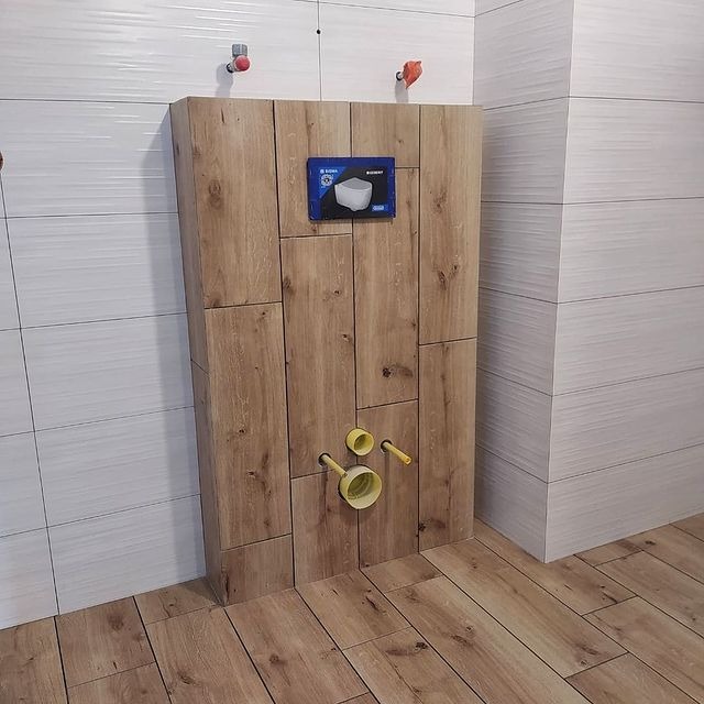 Stelaż podtynkowy WC obudowany płytkami imitującymi drewno, widoczne przyłącza wodne i kanalizacyjne, białe płytki na ścianach i podłodze, niebieska etykieta producenta na stelażu.