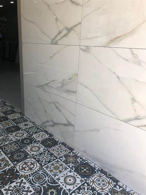 Ściana wyłożona białymi płytami imitującymi marmur z szarymi żyłami, kontrastująca z podłogą pokrytą wzorzystymi, ciemnymi płytkami ceramicznymi.