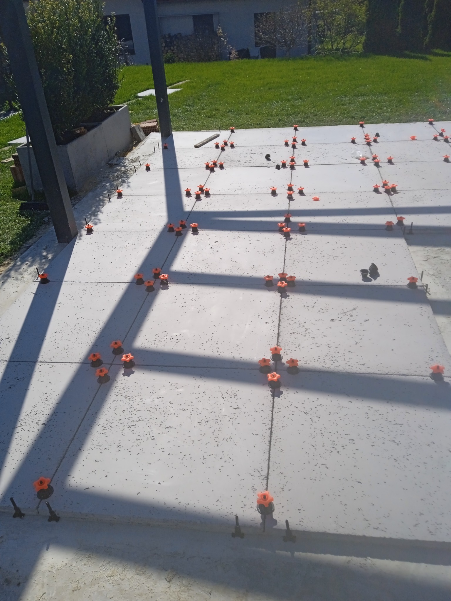 Świeżo ułożone, szare płyty tarasowe z pomarańczowymi elementami poziomującymi i czarnymi klinami, w tle trawnik i budynek.