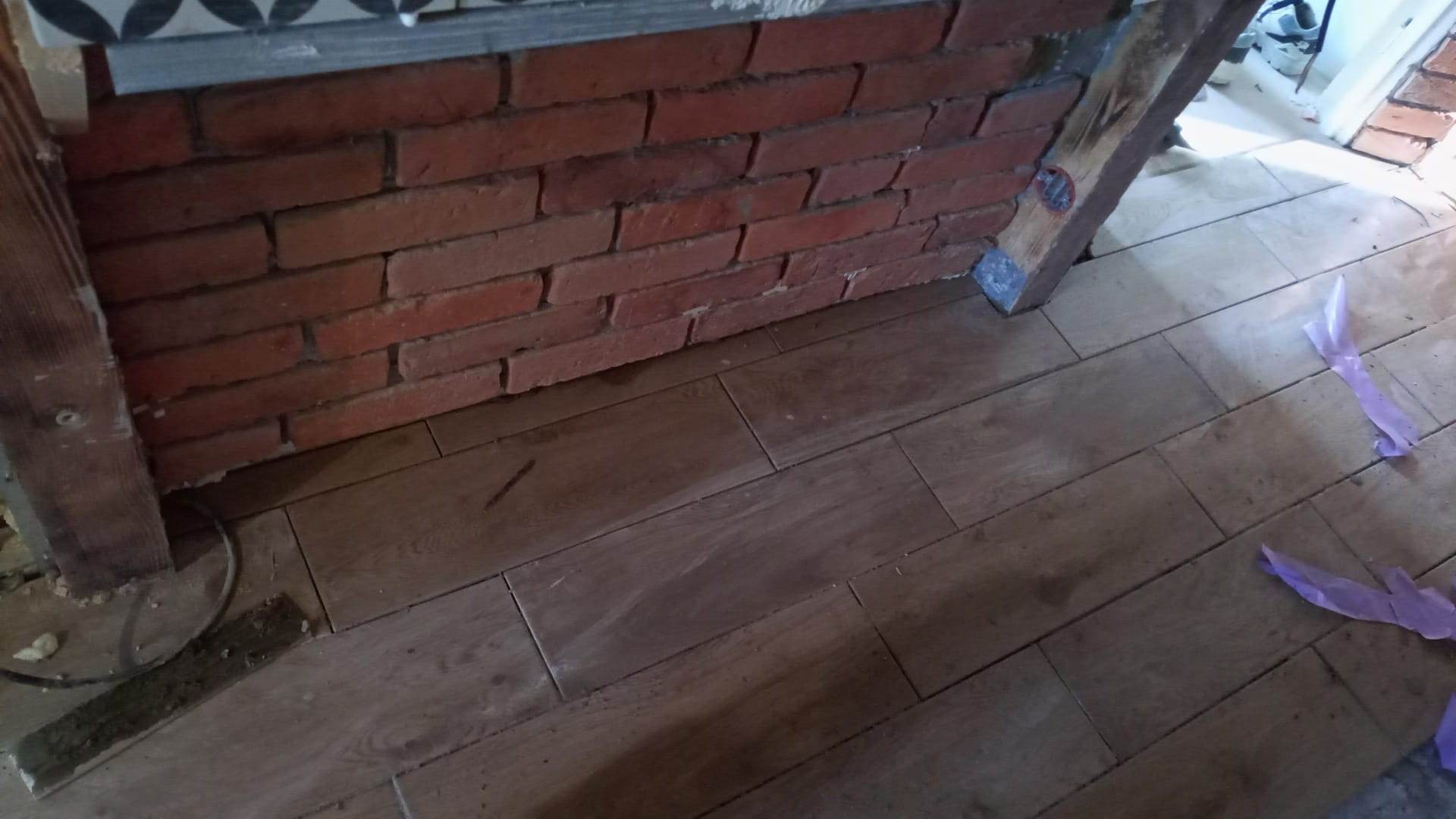 Fragment ściany wyłożonej czerwoną cegłą, z drewnianą ramą i podłogą z paneli imitujących drewno, widoczne ślady prac remontowych i folia ochronna.