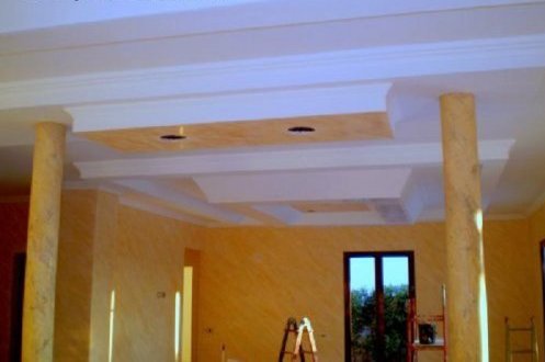 Wnętrze pokoju z kolumnami imitującymi marmur, dekoracyjnym sufitem podwieszanym z oświetleniem punktowym i ścianami pokrytymi farbą strukturalną w odcieniach żółci, drabina malarska w tle.