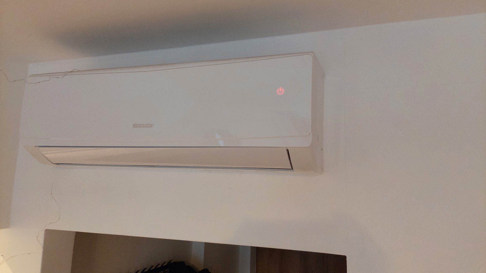 Biały klimatyzator marki Sinclair zamontowany na ścianie, widoczna czerwona dioda zasilania, pod spodem fragment szafki.