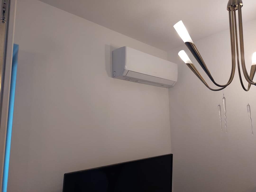 Biały klimatyzator ścienny zamontowany w pokoju z modernistycznym żyrandolem, widoczny fragment czarnego telewizora.