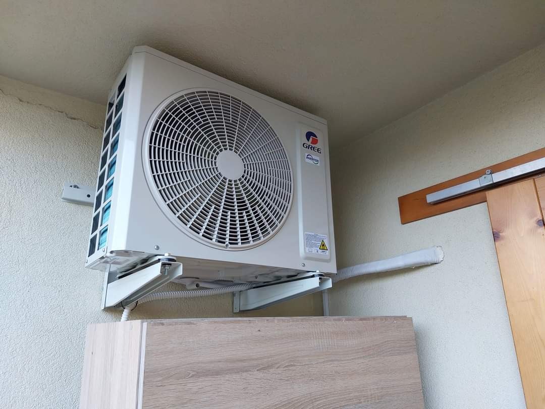 Biała jednostka zewnętrzna klimatyzacji marki Gree zamontowana na wspornikach nad szafką z imitacją drewna, widoczne rury odprowadzające skropliny.