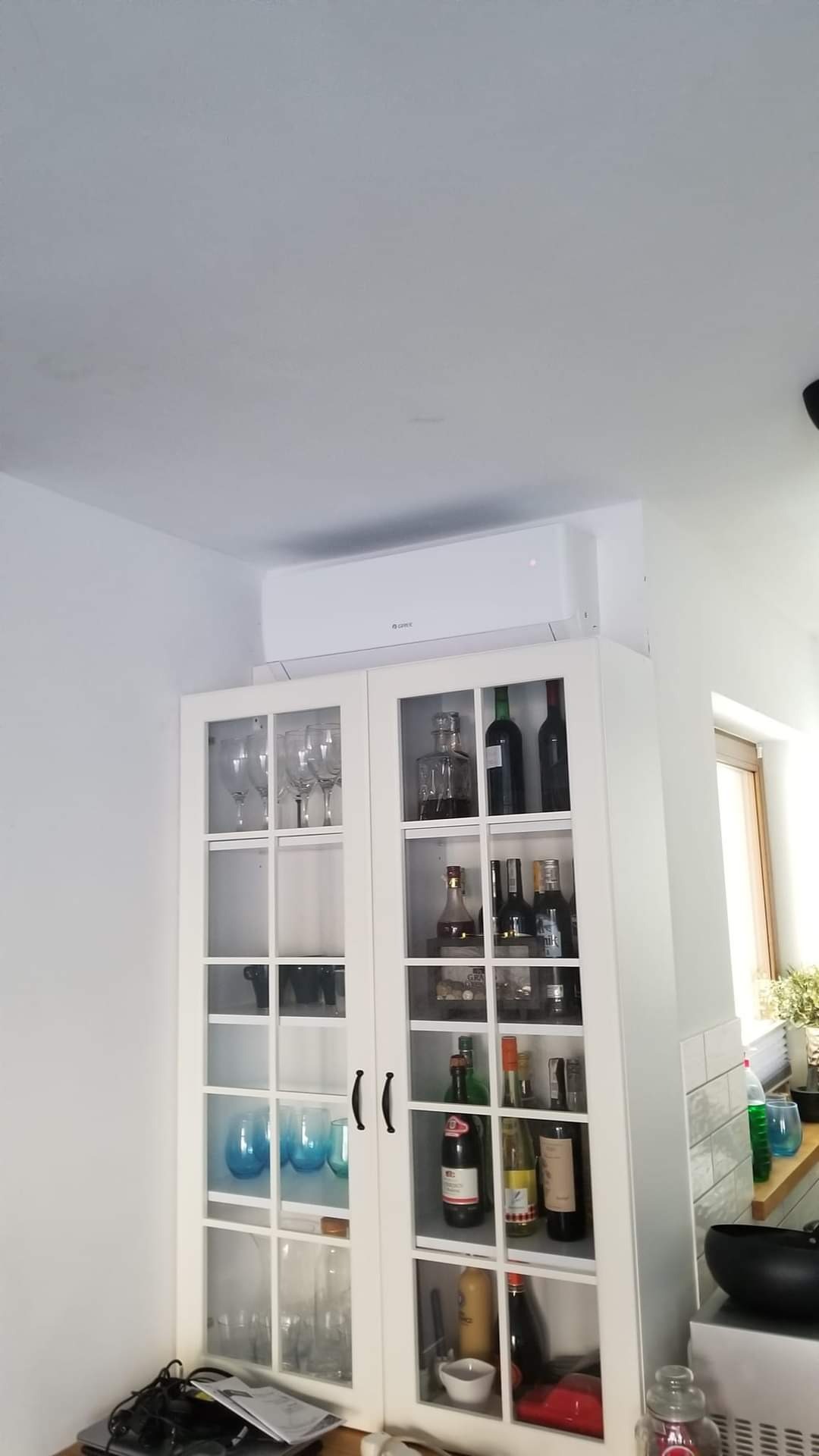 Biały klimatyzator ścienny marki Gree zamontowany nad przeszkloną witryną wypełnioną butelkami i szklankami w jasnym wnętrzu domu.