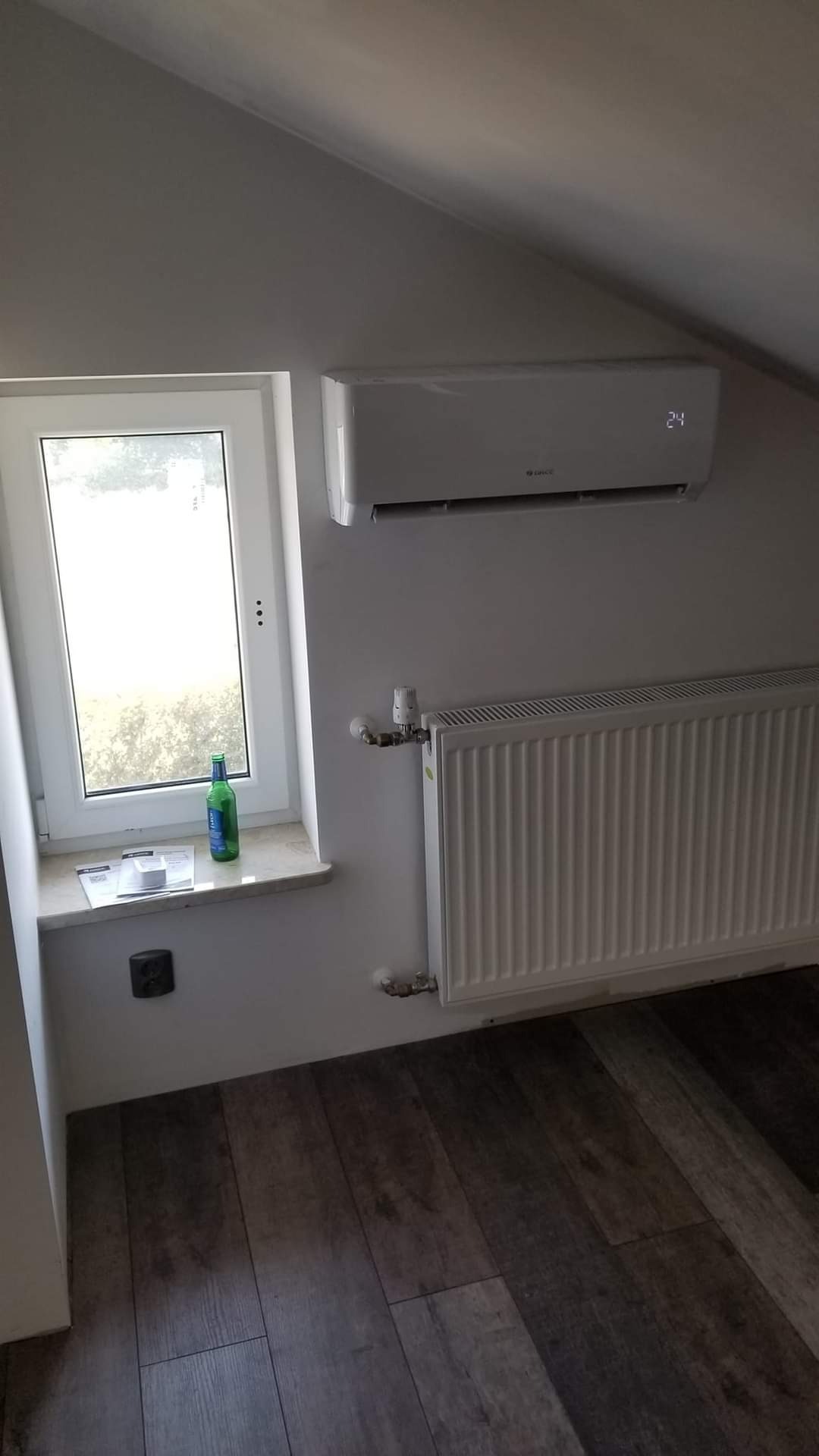 Biały klimatyzator ścienny marki Gree zamontowany w pokoju na poddaszu, obok okna z butelką piwa na parapecie i grzejnika.