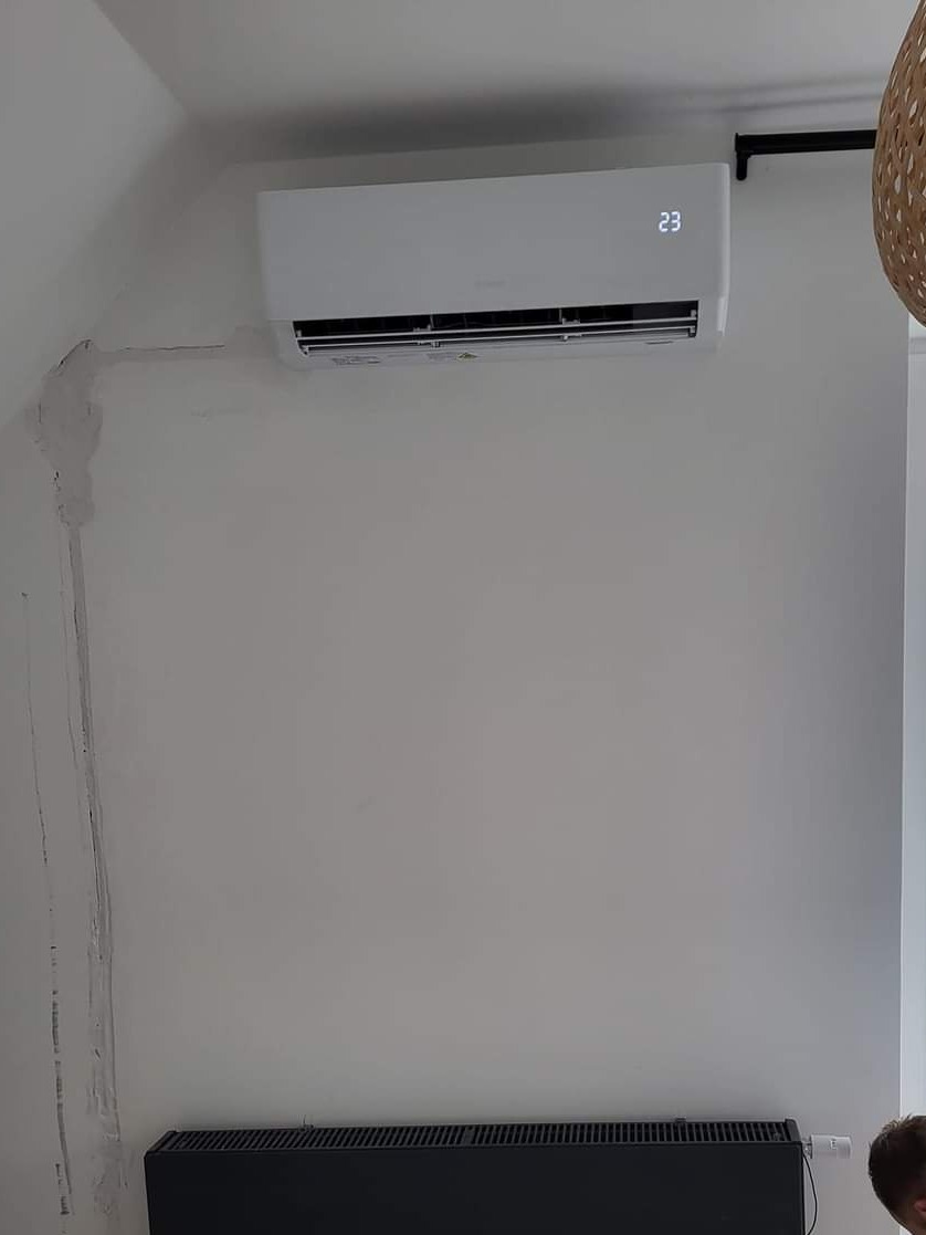 Biała jednostka klimatyzacji na ścianie w pokoju, z widocznymi śladami po instalacji na ścianie, ustawiona temperatura 23 stopnie Celsjusza, grzejnik pod spodem.