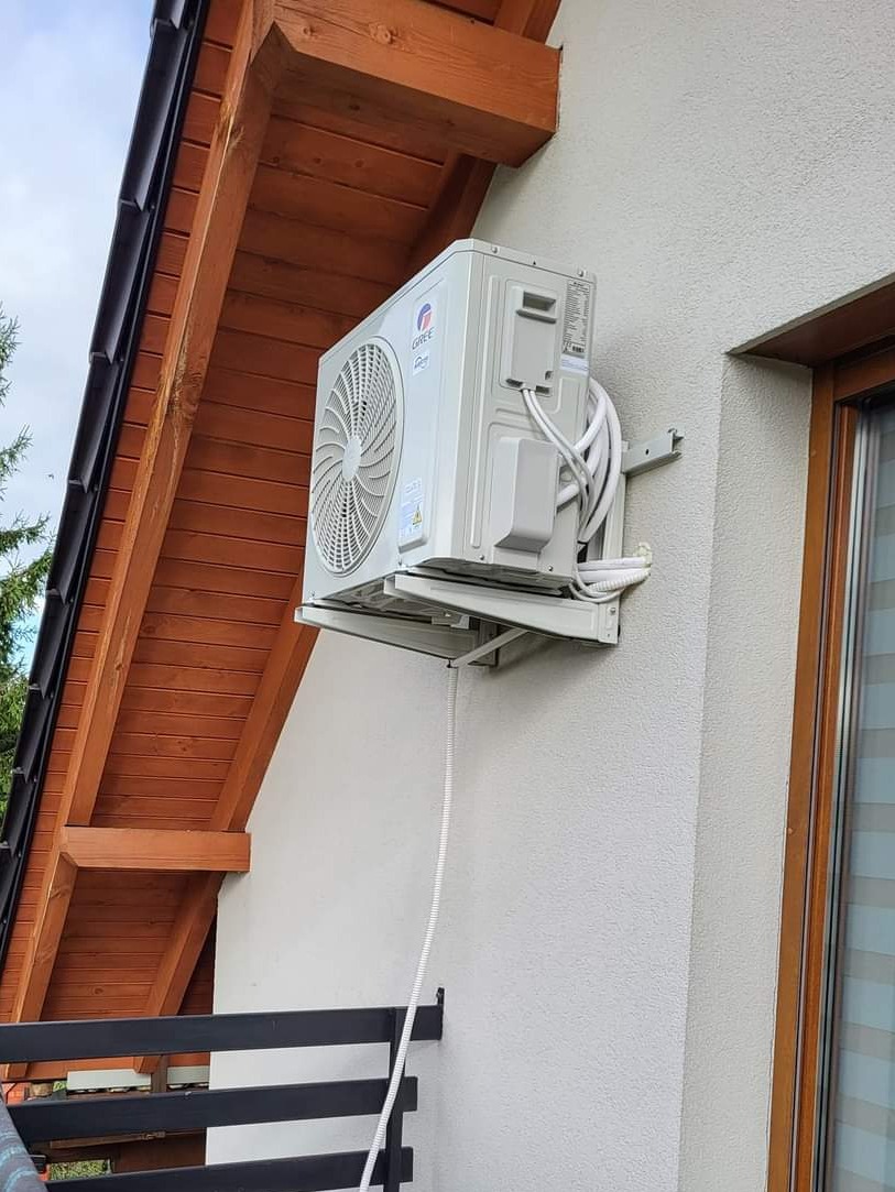 Zewnętrzna jednostka klimatyzacji marki Gree zamontowana na ścianie domu z drewnianym podbitką dachową, widoczna z balkonu.