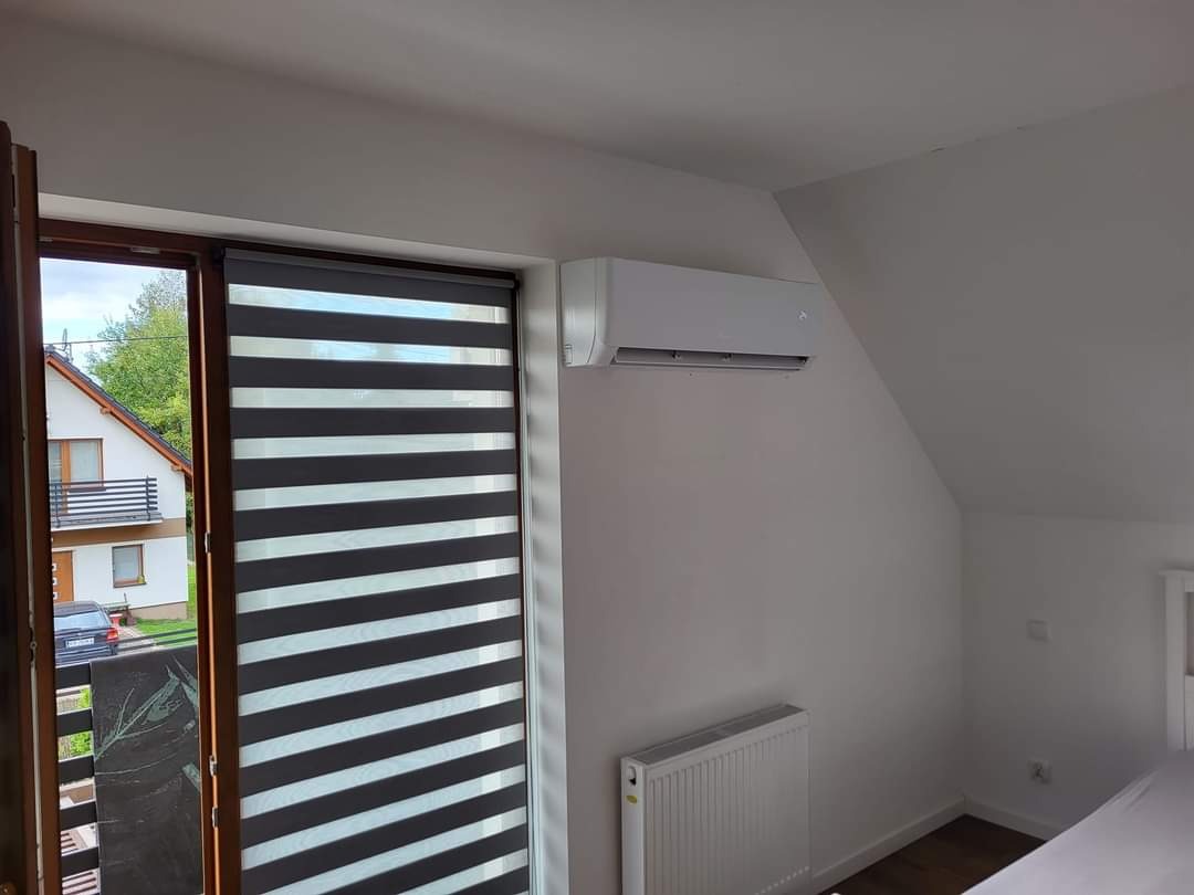 Biała jednostka wewnętrzna klimatyzatora zamontowana na ścianie w pokoju na poddaszu z oknem z żaluzjami dzień-noc i grzejnikiem pod spodem.