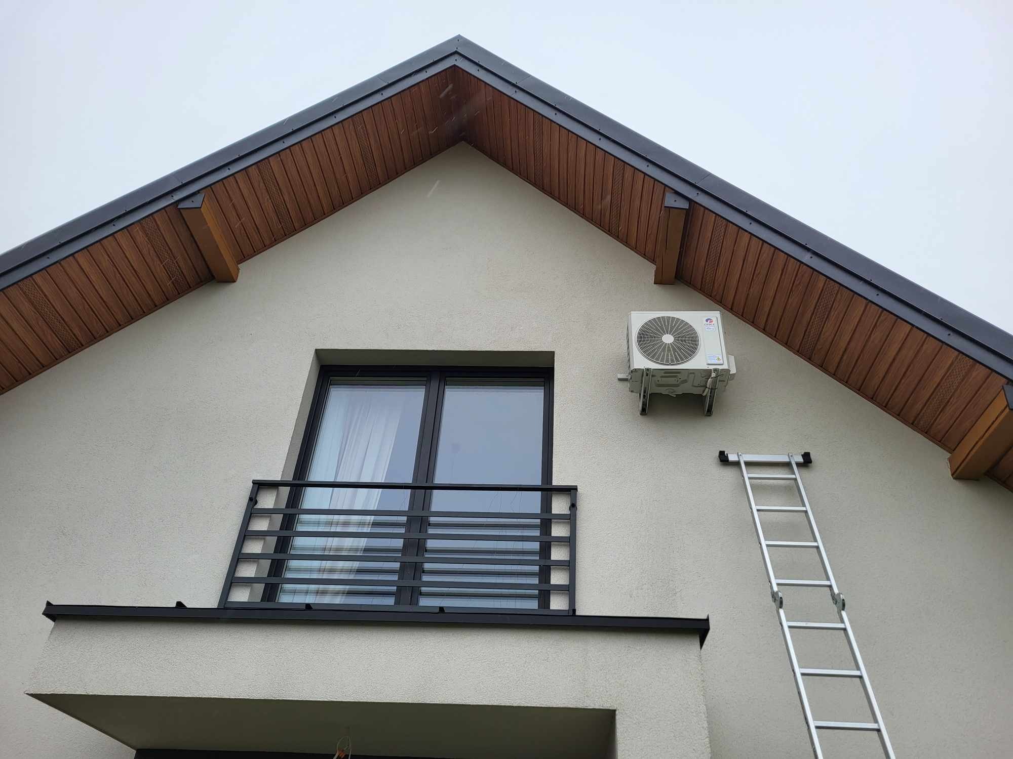 Biała elewacja domu z zamontowanym klimatyzatorem zewnętrznym i przystawioną aluminiową drabiną, widok z dołu.