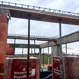 Mateusz Szkopiak Usługi Budowlane - Wznoszenie konstrukcji domu z czerwonej cegły Porotherm, widoczne podpory szalunkowe, zbrojenie stropu i niezabudowany krajobraz w tle.