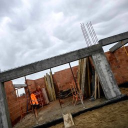 Mateusz Szkopiak Usługi Budowlane - Wznoszenie ścian domu murowanego z betonu i cegły ceramicznej, widoczne zbrojenia pionowe, szalunki, pomarańczowa kurtka robocza i zapas cegieł Wienerberger na placu budowy pod pochmurnym niebem.