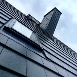 Mateusz Szkopiak Usługi Budowlane - Nowoczesny dach z czarnej dachówki ceramicznej z oknem dachowym i kominem, uchwycony pod kątem w pochmurny dzień, widoczne krople deszczu.