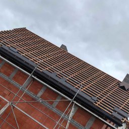 Mateusz Szkopiak Usługi Budowlane - Wznoszenie dachu z dachówki ceramicznej, widoczne łaty i kontrłaty na murze z czerwonej cegły, rusztowanie wzdłuż ściany, pochmurne niebo w tle.