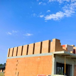 Mateusz Szkopiak Usługi Budowlane - Wznoszenie muru z czerwonej cegły ceramicznej na budowie domu jednorodzinnego, widoczne zbrojenie pionowe i betonowy strop, błękitne niebo w tle.