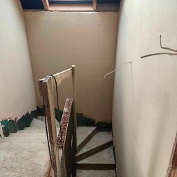 Mateusz Szkopiak Usługi Budowlane - Schody wewnętrzne w trakcie prac wykończeniowych: ściany pokryte świeżo nałożoną gładzią, widoczne drewniane elementy konstrukcyjne schodów i prowizoryczna barierka z cegły, okno dachowe w górnej...