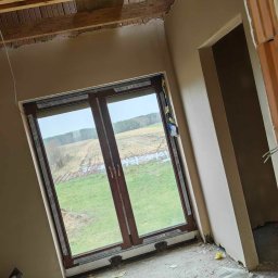 Mateusz Szkopiak Usługi Budowlane - Wnętrze domu w trakcie wykańczania: ściany po nałożeniu gładzi, nowe okno z widokiem na łąkę, surowy strop z drewnianymi belkami, widoczne ślady prac budowlanych.