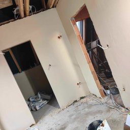 Mateusz Szkopiak Usługi Budowlane - Wnętrze w trakcie prac wykończeniowych: ściany pokryte świeżą warstwą gładzi, widoczne przewody elektryczne wystające ze ścian, surowy strop z drewnianych belek, worki z zaprawą na podłodze...