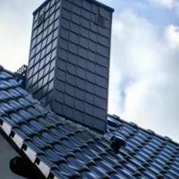 Ciemny, klinkierowy komin z wentylacją, dopasowany kolorystycznie do błyszczącej, ceramicznej dachówki na tle pochmurnego nieba.