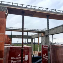 Wznoszenie ścian budynku z czerwonej ceramiki Porotherm, widoczne podpory stropu i zbrojenie, w tle pole i pochmurne niebo.