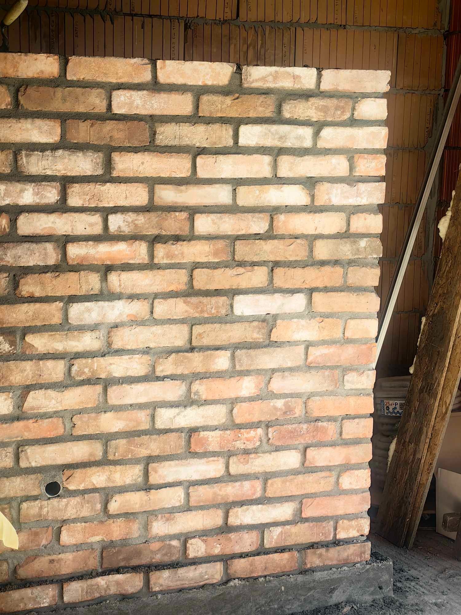 Fragment nowo wymurowanej ściany z cegły klinkierowej o nieregularnych odcieniach, z widocznymi śladami zaprawy i otworem na instalację, na tle ściany z pustaków ceramicznych.