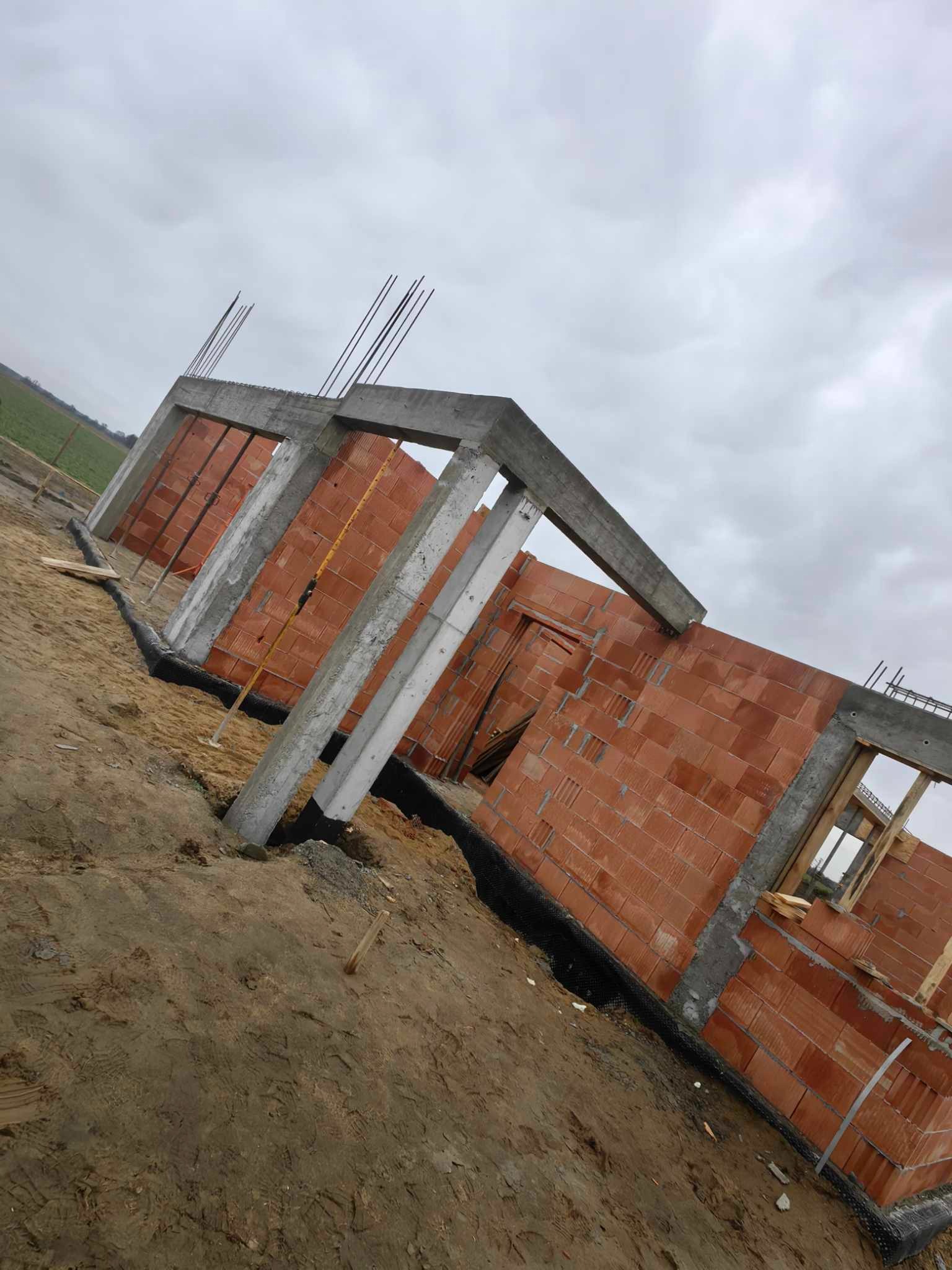 Surowy stan budynku w trakcie wznoszenia ścian z czerwonej cegły, z widocznymi betonowymi słupami i zbrojeniami, na tle pochmurnego nieba i zaoranego pola.