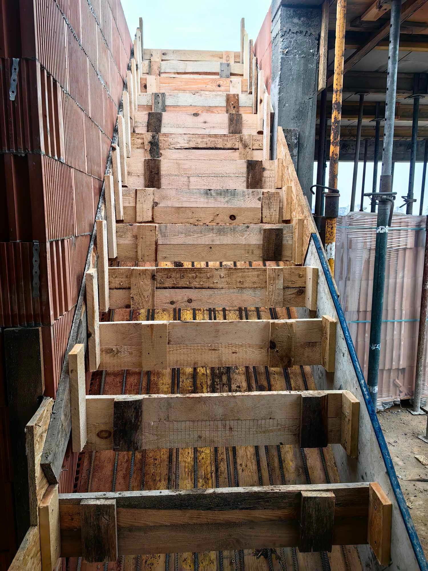 Konstrukcja drewniana szalunku schodów na placu budowy, widoczne zbrojenie i mur z czerwonej cegły dziurawki w tle.