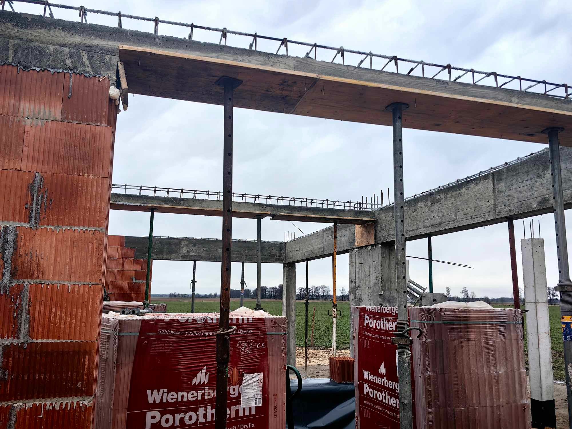 Wznoszenie konstrukcji domu z czerwonej cegły Porotherm, widoczne podpory szalunkowe, zbrojenie stropu i niezabudowany krajobraz w tle.