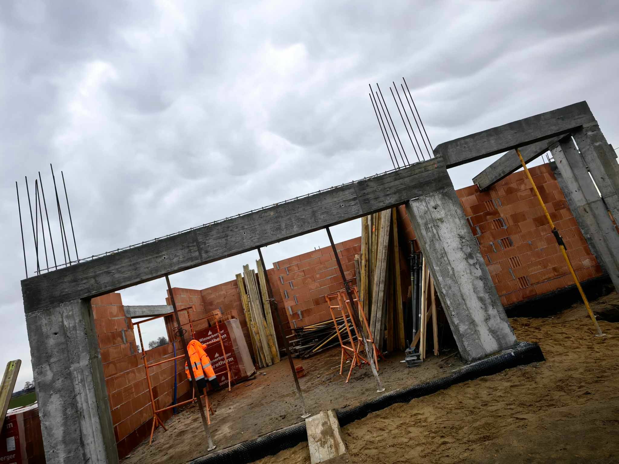 Wznoszenie ścian domu murowanego z betonu i cegły ceramicznej, widoczne zbrojenia pionowe, szalunki, pomarańczowa kurtka robocza i zapas cegieł Wienerberger na placu budowy pod pochmurnym niebem.