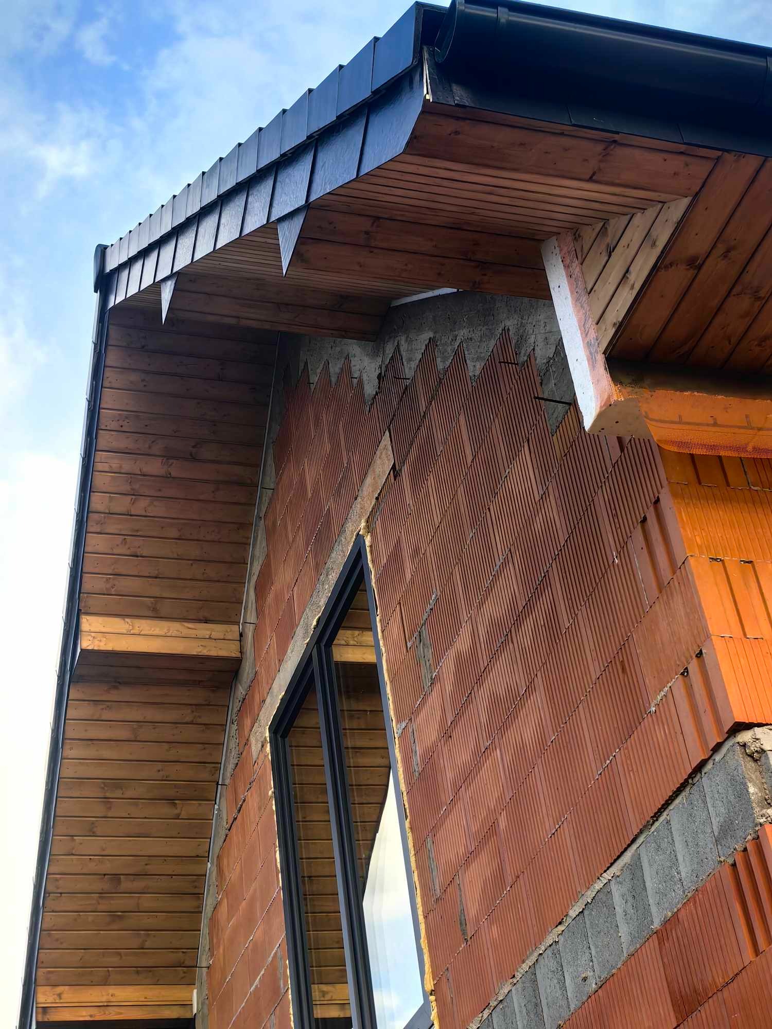 Niski kąt widzenia na budynek w trakcie budowy, widoczne ściany z czerwonej cegły, okno z czarną ramą, drewniana podbitka dachu i fragment niebieskiego nieba.