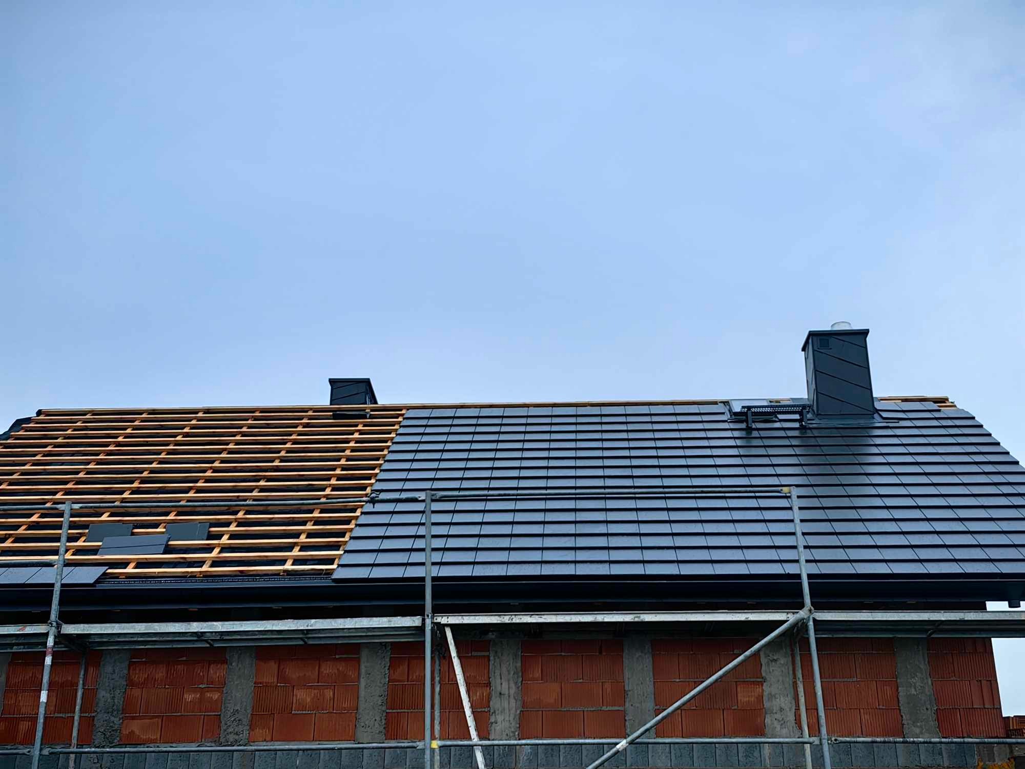 Częściowo pokryty dach domu jednorodzinnego z czarną dachówką, widoczne rusztowania i komin, część dachu z odkrytą konstrukcją drewnianą.