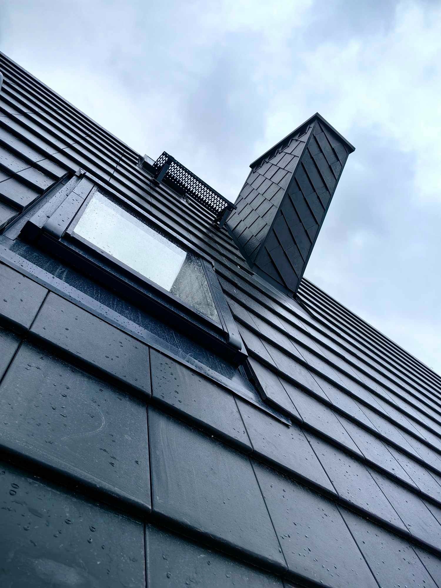 Nowoczesny dach z czarnej dachówki ceramicznej z oknem dachowym i kominem, uchwycony pod kątem w pochmurny dzień, widoczne krople deszczu.