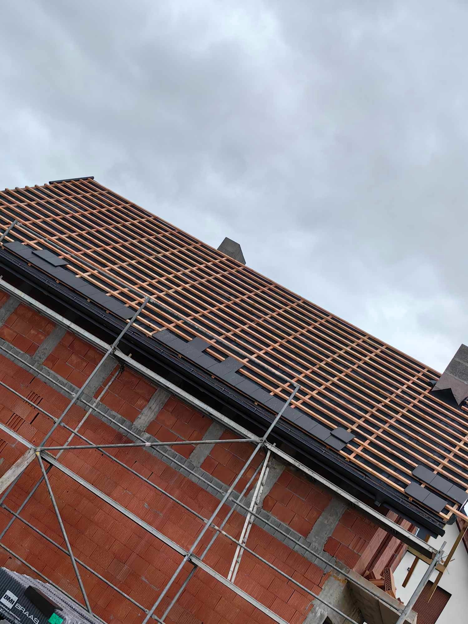 Wznoszenie dachu z dachówki ceramicznej, widoczne łaty i kontrłaty na murze z czerwonej cegły, rusztowanie wzdłuż ściany, pochmurne niebo w tle.