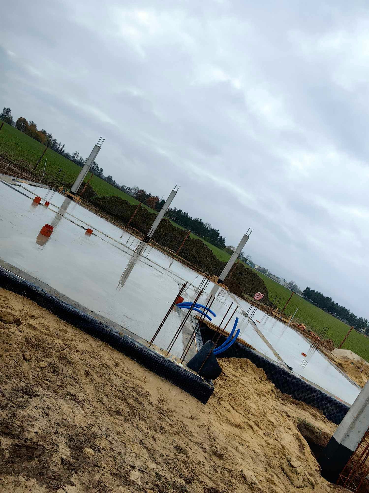 Wylana płyta fundamentowa z wystającymi słupami zbrojeniowymi, widoczne niebieskie rury instalacyjne i czarna folia izolacyjna na obrzeżach, w tle pole i pochmurne niebo.