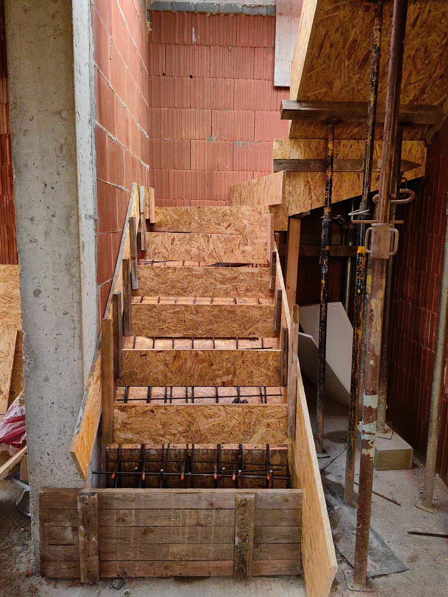 Formowanie schodów z płyty OSB i zbrojenia stalowego w surowym stanie, widoczne podpory i fragment ściany z cegły ceramicznej.