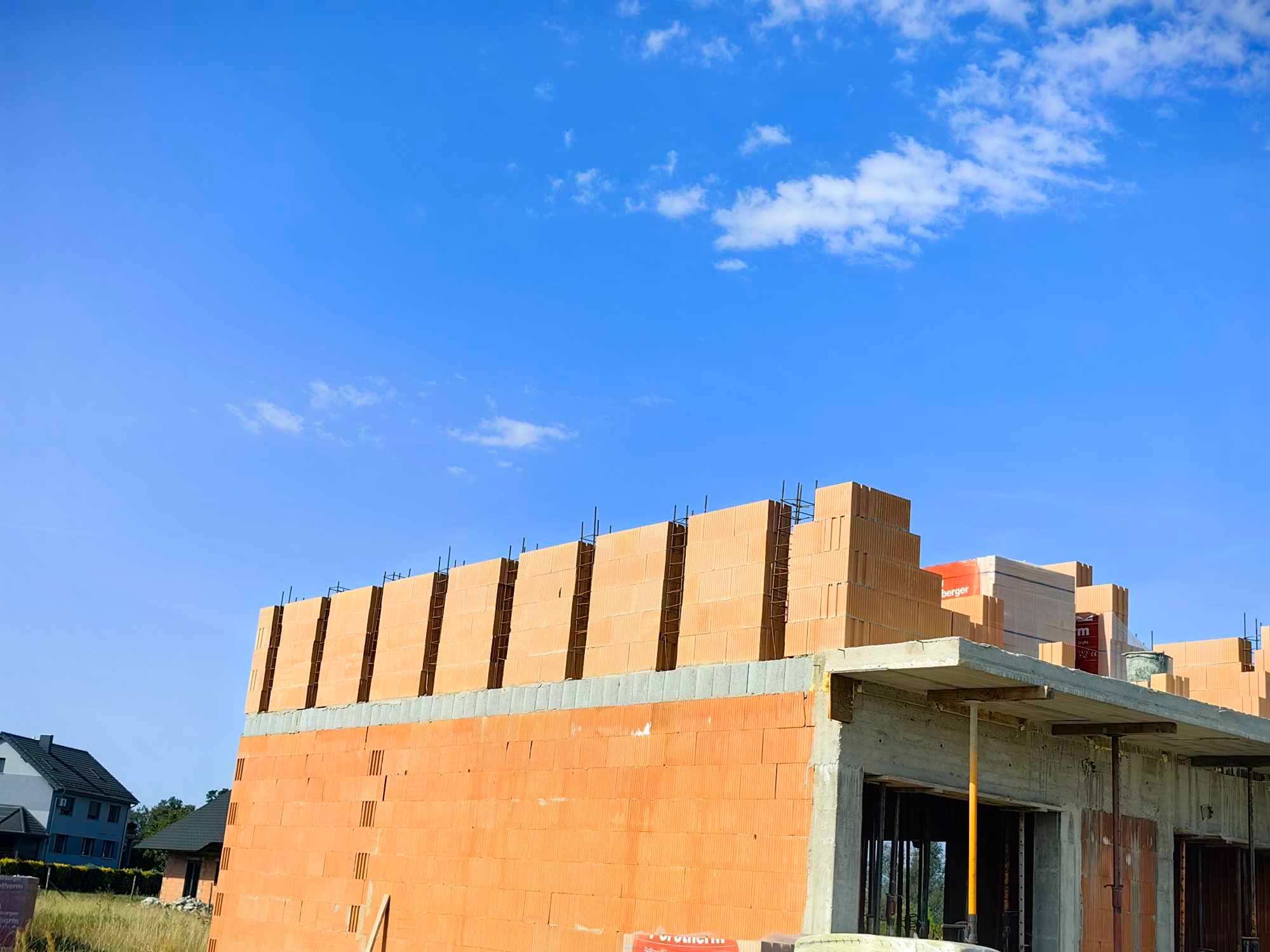 Wznoszenie muru z czerwonej cegły ceramicznej na budowie domu jednorodzinnego, widoczne zbrojenie pionowe i betonowy strop, błękitne niebo w tle.